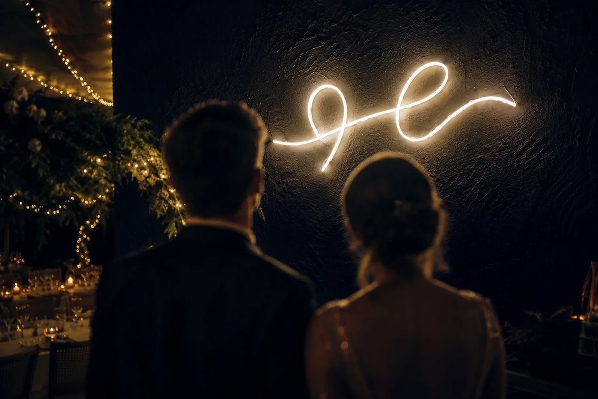 Néon LED blanc chaud en écriture cursive sur le mur d'une réception de mariage, deux invités de dos